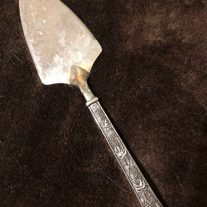 Puede incluir: Un servidor de pastel de plata vintage con una hoja ancha y triangular y un mango decorativo. El mango presenta un diseño en relieve de racimos de uvas y hojas. El servidor se encuentra sobre un fondo marrón oscuro texturizado.