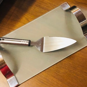 Könnte beinhalten: Ein rechteckiges Serviertablett mit hellgrüner Oberfläche und gebogenen silbernen Metallkanten. Ein Kuchenheber aus Edelstahl mit schwarzem Griff liegt auf dem Tablett.