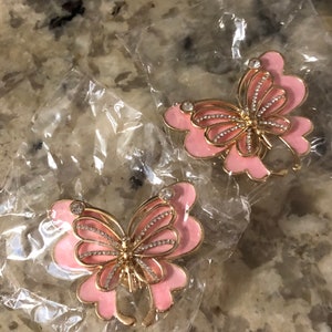 May include: Two pink and gold butterfly brooches with rhinestone accents are displayed in clear plastic packaging. The brooches have a gold outline and are in the shape of a butterfly with pink enamel wings and a rhinestone-studded body.
