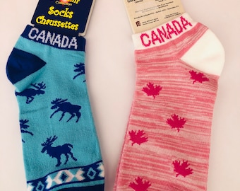 May include: Two souvenir socks with tags. One is light blue with moose silhouettes and the word "CANADA". The other is pink with maple leaf designs and "CANADA" in white letters. Both socks are souvenir items.