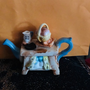 May include: A ceramic teapot with a light blue handle and spout. The teapot is decorated with a small table on top, holding a basket of peaches, a small creamer, and a black object. The teapot has a rustic, cottagecore aesthetic.