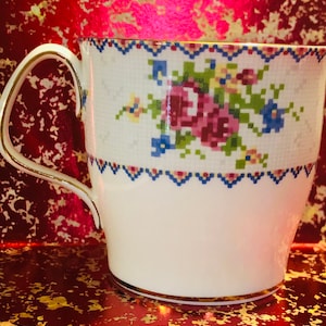 Peut inclure: Tasse blanche avec un motif floral rose, bleu et vert. La tasse a une anse et une bordure dorées. Le motif floral est un design pixélisé. La tasse est sur une surface rouge.