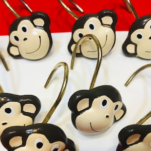 May include: A collection of shower curtain hooks featuring smiling monkey faces. The monkey faces are painted with a cream-colored face, black hair, and a gold-colored hook. The hooks are arranged in rows against a white and red background.