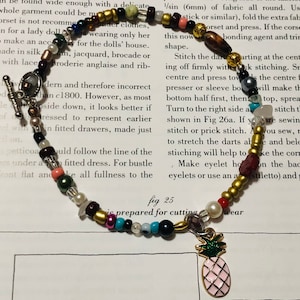 Pode incluir: Uma pulseira de contas com uma variedade de contas coloridas, incluindo dourado, preto, rosa e turquesa. Um pingente de abacaxi dourado com esmalte rosa pende da pulseira. A pulseira é exibida em uma página de livro vintage.