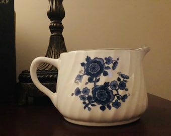 May include: White ceramic creamer with a blue floral design. The creamer has a curved handle and a spout for easy pouring. The floral pattern features detailed blue flowers and leaves. The creamer has a silver trim around the top edge.
