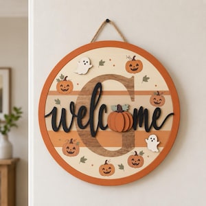 May include: A round wooden wall sign featuring a large letter G, the word welcome in black script, and decorative elements including orange pumpkins, white ghosts, and small green leaves against a background of horizontal tan and orange stripes.