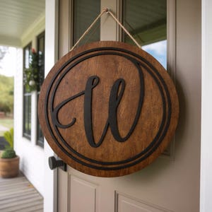 May include: A circular wooden door sign featuring a dark brown stained finish and a black, script-style letter W centered within a thin black circular border. The sign is suspended by a light-colored jute rope attached to the top edge.