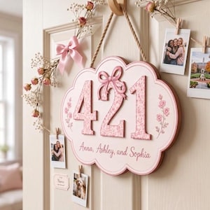 May include: A scalloped wooden wall sign featuring the number 421 in a pink floral pattern, topped with a pink bow. Below the number, the text reads Anna, Ashley, and Sophia. The sign hangs from a rope and is surrounded by dried flowers and photos.