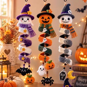 Könnte beinhalten: Drei hängende Halloween-Dekorationen aus Häkelarbeit, bestehend aus einem Geist mit violettem Hut, einem Kürbis mit schwarzem Hut und einem Totenkopf mit dunkelblauem Hut. Jedes Ornament hat einen langen, spiralförmigen Körper mit kleinen Motiven.