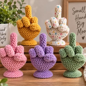 Puede incluir: Cinco esculturas de manos tejidas a ganchillo en amarillo, blanco con motas multicolores, rosa, morado y verde. Cada mano está colocada con el dedo medio extendido hacia arriba. Las figuras tienen textura de lana y bases circulares.