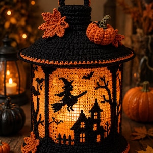 May include: A black and orange crocheted lantern featuring a silhouette of a witch on a broomstick flying over a house. The lantern is decorated with crocheted maple leaves and a small pumpkin on top, with a warm orange light glowing from within.
