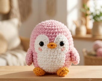 Puede incluir: Un juguete de peluche de pingüino tejido a ganchillo y redondeado, con un cuerpo de color rosa suave, una barriga y una mancha facial blancas, dos ojos de plástico negro, un pequeño pico naranja y dos pies naranjas. La textura del hilo es visible.