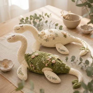 May include: Two crocheted dinosaur plush toys with long necks and tails. One is cream-colored with white floral embroidery, and the other is sage green with pink and white floral embroidery. Both have closed eyes, pink cheeks, and leaf-like tail fins.