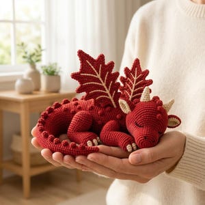 Peut inclure: Un petit dragon en crochet fait main de couleur rouge foncé. Le dragon présente de grandes ailes en forme de feuille avec des détails de nervures claires, de petites cornes sur la tête et une longue queue texturée avec des pointes arrondies.