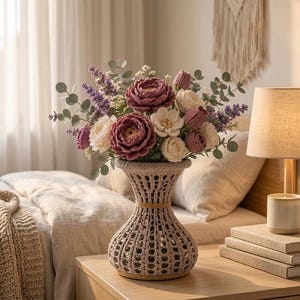 May include: A beige crochet vase with a flared neck and a wide base, containing a bouquet of crocheted flowers in shades of mauve, cream, and purple, accented with green eucalyptus leaves and small white blossoms, arranged in a dense, rounded cluster.