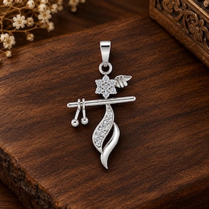 Könnte beinhalten: Ein silberfarbener Anhänger in Form einer Flöte mit einem horizontalen Stab, zwei kleinen hängenden Kugeln, einem sechsblättrigen Blumenmotiv und einem geschwungenen blattartigen Element. Die Blumen- und Blattelemente sind mit kleinen Steinen besetzt.