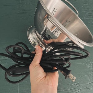 May include: A silver colander with a black electrical cord and plug. The colander has a handle and a base with holes.