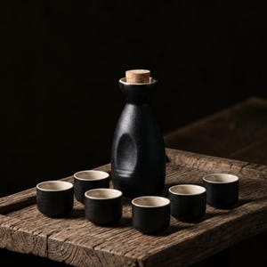 Op de afbeelding: Een sake-set van zwarte keramiek bestaande uit een hoge fles met een kurken stop en zes kleine bijpassende kopjes. Elk stuk heeft een getextureerde matzwarte buitenkant en een gladde, licht crèmekleurige binnenkant voor contrast.