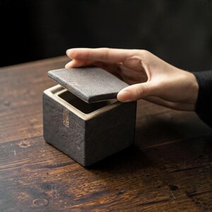 May include: A square, dark gray ceramic container with a textured, stone-like exterior finish and a matching removable lid. The interior of the box is light-colored, and a small, rectangular metallic accent is visible on the front side.