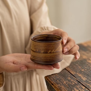 May include: A small, cylindrical ceramic cup featuring a textured, earthy brown finish with horizontal ridges circling the exterior. The vessel has a wide, open top and a slightly tapered base, showcasing a rustic, hand-thrown appearance.