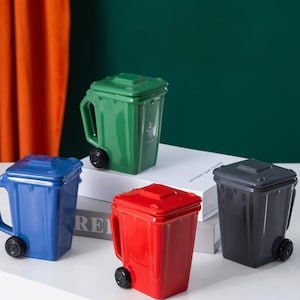 Puede incluir: Cuatro tazas de cerámica con forma de cubos de basura en miniatura con tapas y pequeñas ruedas negras. Las tazas son de color verde, azul, rojo y gris oscuro. Cada taza cuenta con un asa lateral y un diseño de cuerpo rectangular.