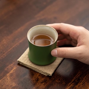 Può includere: Una tazza cilindrica in ceramica con esterno verde oliva opaco e interno beige chiaro, riempita con un liquido marrone. La tazza poggia su un sottobicchiere quadrato, testurizzato e color beige chiaro, dal design semplice e funzionale.
