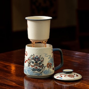 May include: A ceramic tea mug with a removable infuser basket and a matching lid. The mug features a textured off-white finish with a colourful graphic of a stylised lion head surrounded by blue clouds and red ribbons. It has a dark blue handle.