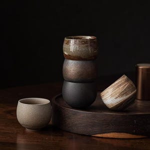 May include: A collection of five ceramic cups with varied textures and finishes. Three cups are stacked vertically, one sits upright on the left, and one rests tilted on its side. The colors include mottled brown, metallic bronze, matte black, and beige.