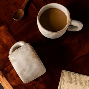 May include: Two off-white ceramic mugs with a textured, uneven finish. One mug is upright and filled with brown coffee, while the other lies on its side. A small wooden spoon and a piece of printed newspaper are visible nearby on the dark surface.