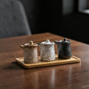 Puede incluir: Tres pequeños tarros de cerámica para condimentos con tapa, cada uno con un esmalte moteado único en marrón, blanco y negro. Los tarros están dispuestos en fila sobre una bandeja rectangular de madera clara, cada uno con un pequeño pico vertedor.