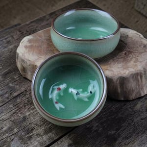 Könnte beinhalten: Zwei runde Keramik-Teeschalen mit einer hellen mintgrünen Glasur und einer subtilen Rissoptik. Das Innere der vorderen Schale zeigt zwei kleine, erhabene weiße Koi-Fischfiguren mit roten und schwarzen Markierungen am Boden.