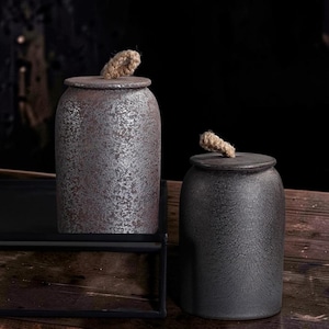 May include: Two cylindrical ceramic canisters with lids, each topped with a small jute rope handle. The taller canister features a mottled silver and dark brown finish, while the shorter canister has a textured, dark charcoal gray surface.