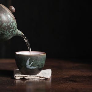 May include: A ceramic tea cup with a dark green and brown mottled glaze featuring a white crane illustration. Tea is being poured into the cup from a matching teapot. The cup rests on a small, folded, light-colored fabric square coaster.