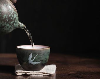 Puede incluir: Una taza de té de cerámica con esmalte moteado en verde oscuro y marrón, con una ilustración de una grulla blanca. Se vierte té en la taza desde una tetera a juego. La taza descansa sobre un pequeño cuadrado de tela clara doblado.