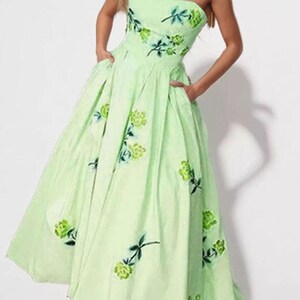May include: A strapless, floor-length light green dress featuring a sweetheart neckline and a full, pleated skirt with side pockets. The fabric is adorned with a scattered pattern of embroidered green roses with dark green stems and leaves.