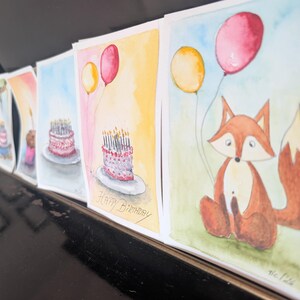 May include: A series of watercolor greeting cards lined up, featuring illustrations of a fox holding yellow and red balloons, and birthday cakes with lit candles. One card clearly displays the handwritten text Happy Birthday in black ink.