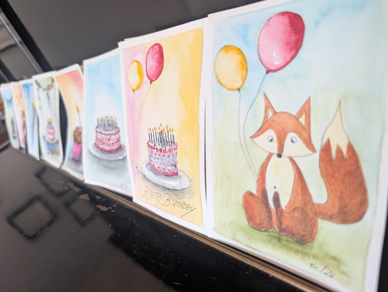 May include: A series of watercolor greeting cards lined up, featuring illustrations of a fox holding yellow and red balloons, and birthday cakes with lit candles. One card clearly displays the handwritten text Happy Birthday in black ink.