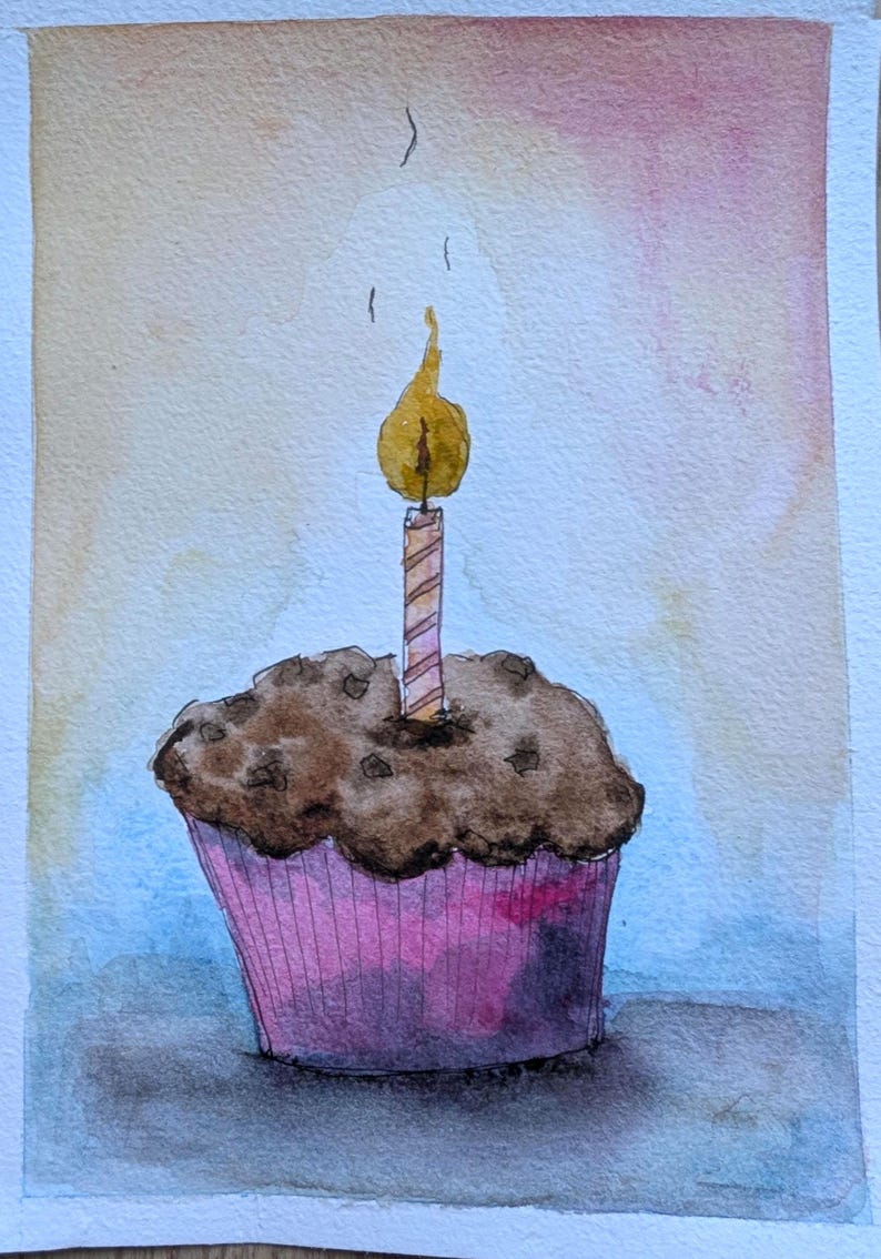 May include: A watercolor painting of a chocolate muffin in a pink paper liner with a single lit birthday candle on top. The candle has a yellow flame and a striped pattern. The background features a soft gradient of light orange and blue tones.
