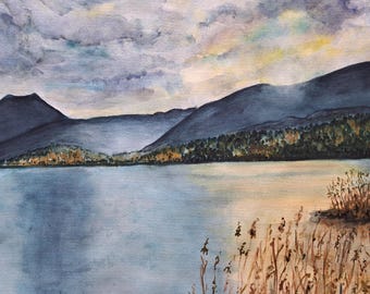 May include: A watercolor painting depicting a serene lake landscape with dark blue mountains in the background under a cloudy sky. The foreground features tall, dry golden-brown reeds along the water's edge, reflecting soft light across the surface.