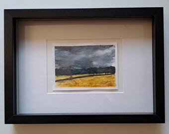 May include: A framed watercolor painting depicting a landscape with a golden yellow field under a dark, stormy gray sky. The artwork features a thin horizontal line representing a fence or power line and is encased in a simple black frame with a white mat.