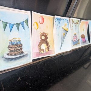 May include: A series of watercolor greeting cards featuring various celebratory illustrations, including a chocolate cake with blue candles, a brown teddy bear with yellow and red balloons, and a school cone with the number one printed on it.