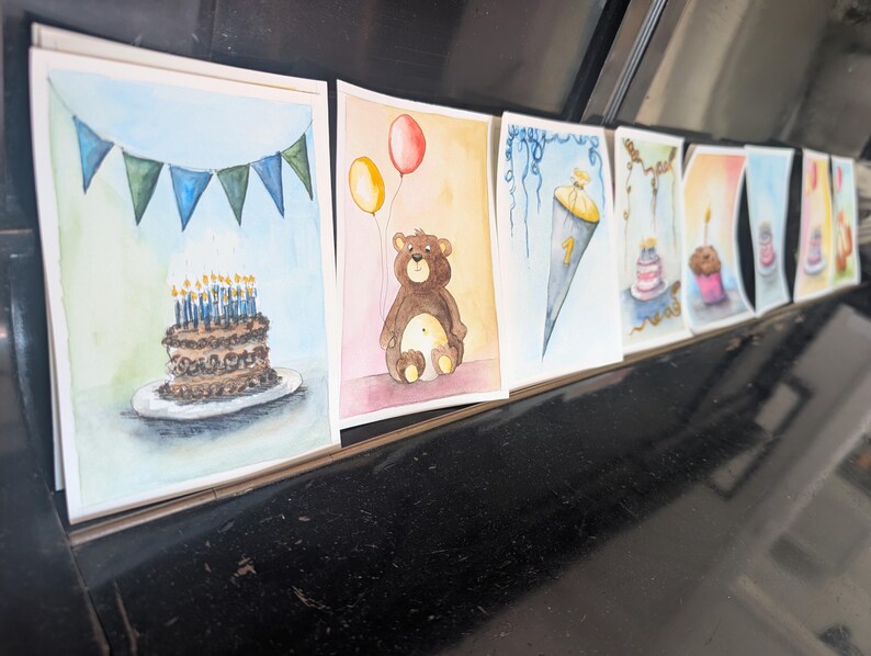 May include: A series of watercolor greeting cards featuring various celebratory illustrations, including a chocolate cake with blue candles, a brown teddy bear with yellow and red balloons, and a school cone with the number one printed on it.