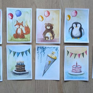 May include: A collection of fourteen rectangular watercolor paintings featuring birthday themes, including cakes with candles, a fox, a bear, a penguin, balloons, bunting banners, and a school cone, all arranged in two rows on a wooden surface.