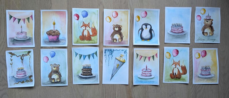 May include: A collection of fourteen rectangular watercolor paintings featuring birthday themes, including cakes with candles, a fox, a bear, a penguin, balloons, bunting banners, and a school cone, all arranged in two rows on a wooden surface.