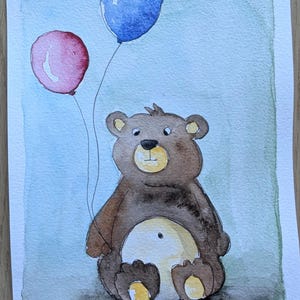 May include: A watercolor painting on paper depicting a brown bear sitting down and holding two balloons, one red and one blue. The bear has a light tan belly and muzzle. The background is a soft, blended wash of light blue and green. Signed 'nic '26'.