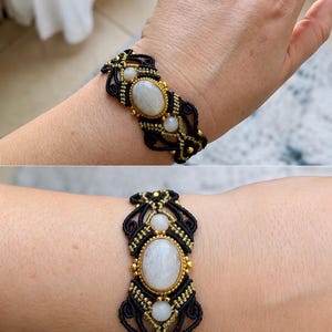 May include: A macrame bracelet featuring a large oval moonstone cabochon at the centre, flanked by two smaller round moonstone beads. The design incorporates black and gold cord woven into intricate patterns with small gold metallic accents.