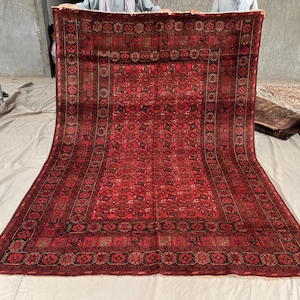 Op de afbeelding: Een groot rechthoekig vloerkleed met een dieprode achtergrond en ingewikkelde zwarte en donkerbruine geometrische patronen. Het kleed heeft een brede, gedetailleerde rand met herhalende florale en tribale motieven, afgewerkt met korte franjes.