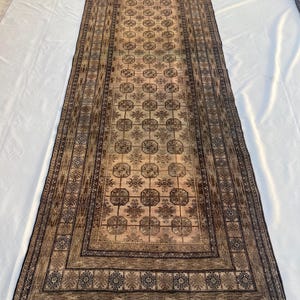 May include: A long rectangular runner rug featuring a beige central field with a repeating grid of dark brown octagonal motifs and floral elements. The rug is framed by multiple decorative borders in shades of brown, tan, and black with fringe.