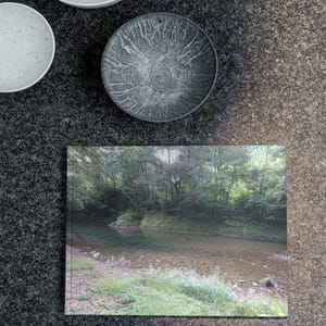 Pode incluir: Um livro retangular com uma fotografia de paisagem de um rio a fluir através de uma floresta, acompanhado por duas taças brancas salpicadas e uma taça cinzenta escura com um padrão ramificado e texturado na superfície interior.