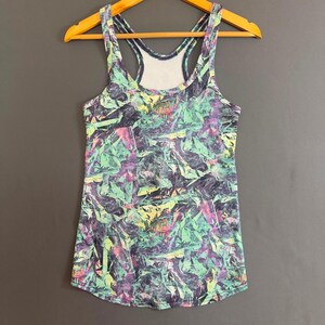 May include: A sleeveless racerback tank top featuring an abstract, multi-colored print with shades of dark purple, mint green, and bright pink. The garment has a scoop neckline and is displayed on a wooden hanger against a plain gray background.